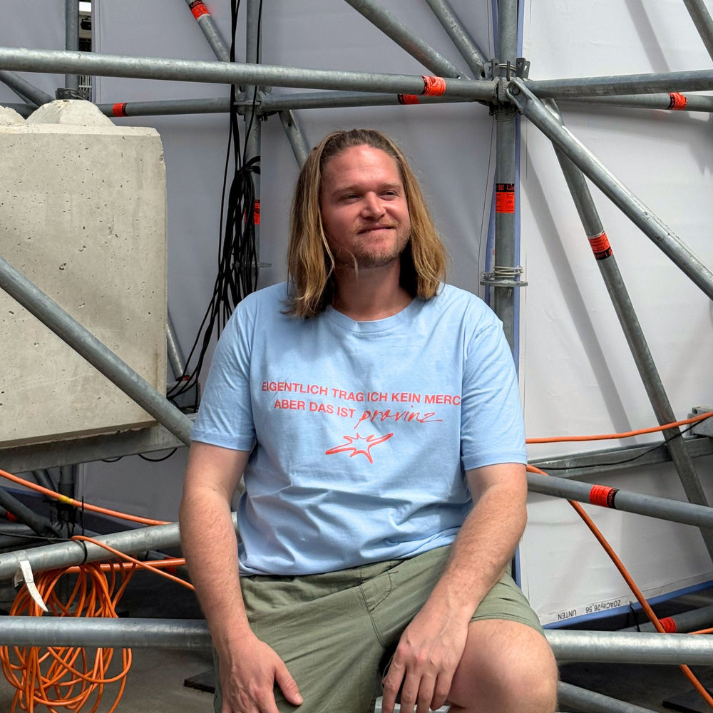 Ein Mann mit schulterlangen blonden Haaren sitzt vor einem Metall-Bühnengerüst und trägt das hellblaue T-Shirt der Band Provinz mit dem roten Spruch-Aufdruck auf der Brust.