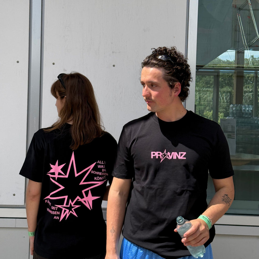 Zwei Personen tragen das schwarze Provinz T-Shirt im Freien: Links zeigt eine Person die Rückseite mit der rosa Sternengrafik und dem Spruch, rechts steht ein Mann im Vordergrund und präsentiert die Brustansicht mit rosa Logo.