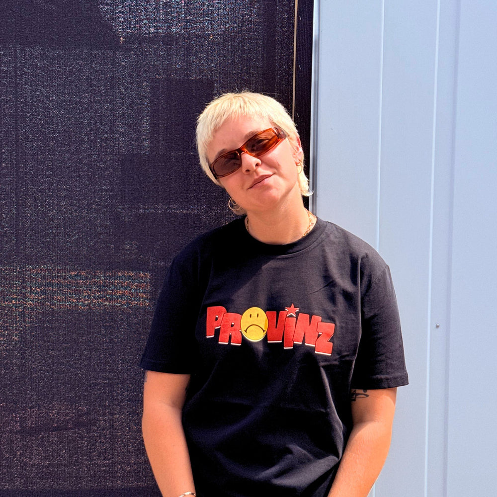 Eine Person mit blondem Kurzhaarschnitt und braun getönter Sonnenbrille im Portrait, die das schwarze T-Shirt der Band Provinz mit dem roten Schriftzug und gelbem Sad-Smiley auf der Brust trägt.