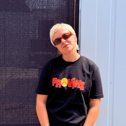 Eine Person mit blondem Kurzhaarschnitt und braun getönter Sonnenbrille im Portrait, die das schwarze T-Shirt der Band Provinz mit dem roten Schriftzug und gelbem Sad-Smiley auf der Brust trägt.