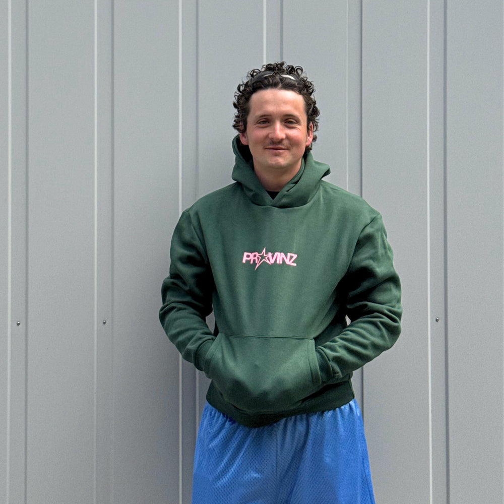Ein Mann steht vor einer grauen Metallwand, Er trägt den dunkelgrünen Hoodie der Band Provinz mit dem rosa Stern-Logo auf der Brust und hat seine Hände in die Kängurutasche gesteckt. Dazu trägt er eine blaue Sporthose.