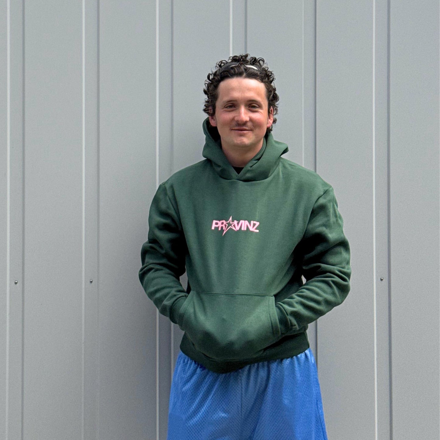 Ein Mann steht vor einer grauen Metallwand, Er trägt den dunkelgrünen Hoodie der Band Provinz mit dem rosa Stern-Logo auf der Brust und hat seine Hände in die Kängurutasche gesteckt. Dazu trägt er eine blaue Sporthose.