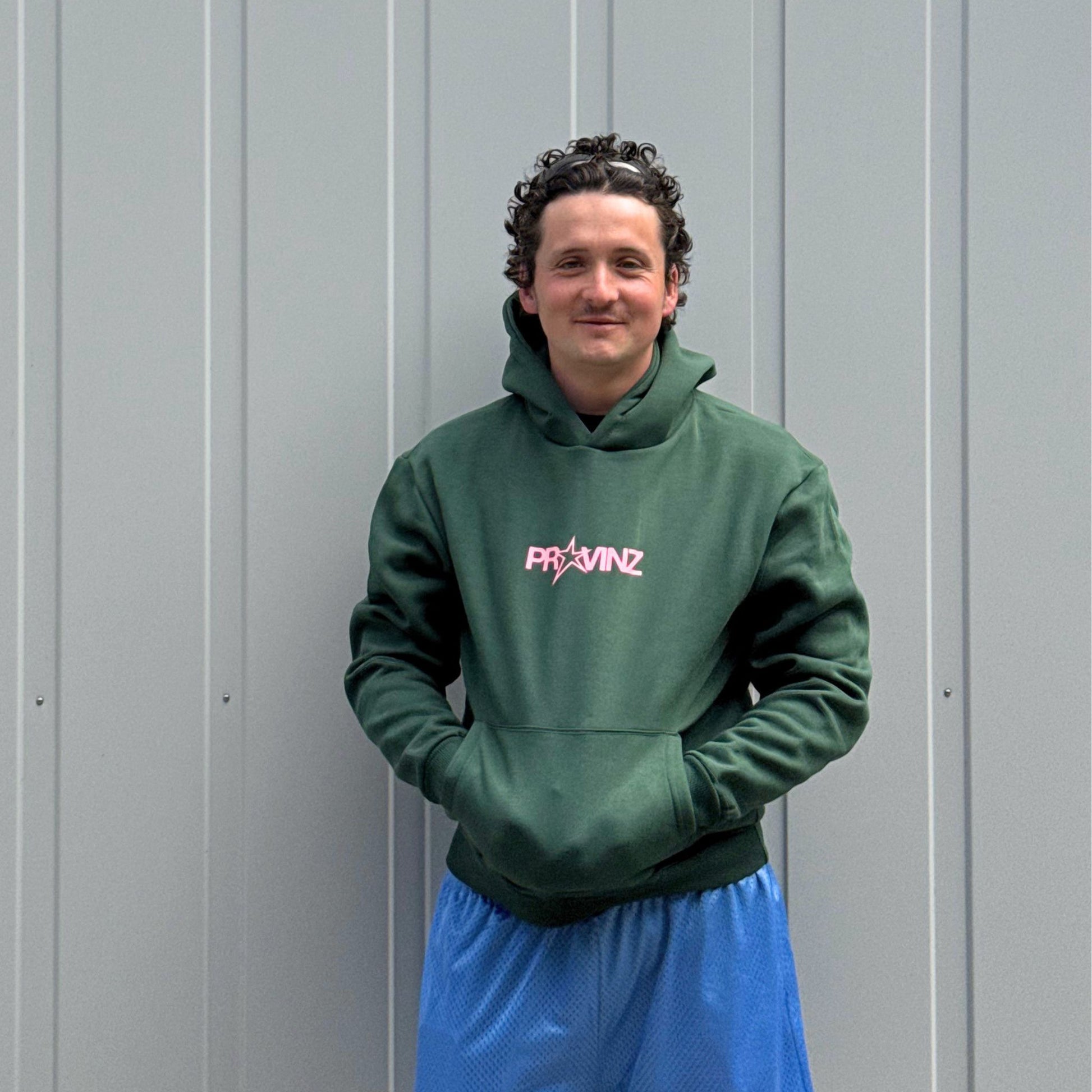 Ein Mann steht vor einer grauen Metallwand, Er trägt den dunkelgrünen Hoodie der Band Provinz mit dem rosa Stern-Logo auf der Brust und hat seine Hände in die Kängurutasche gesteckt. Dazu trägt er eine blaue Sporthose.