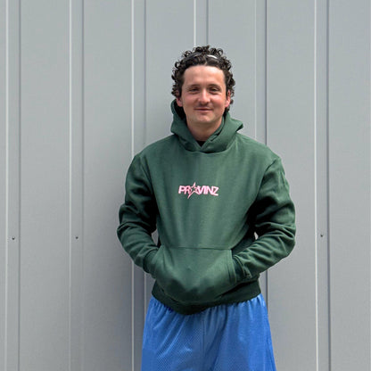 Ein Mann steht vor einer grauen Metallwand, Er trägt den dunkelgrünen Hoodie der Band Provinz mit dem rosa Stern-Logo auf der Brust und hat seine Hände in die Kängurutasche gesteckt. Dazu trägt er eine blaue Sporthose.